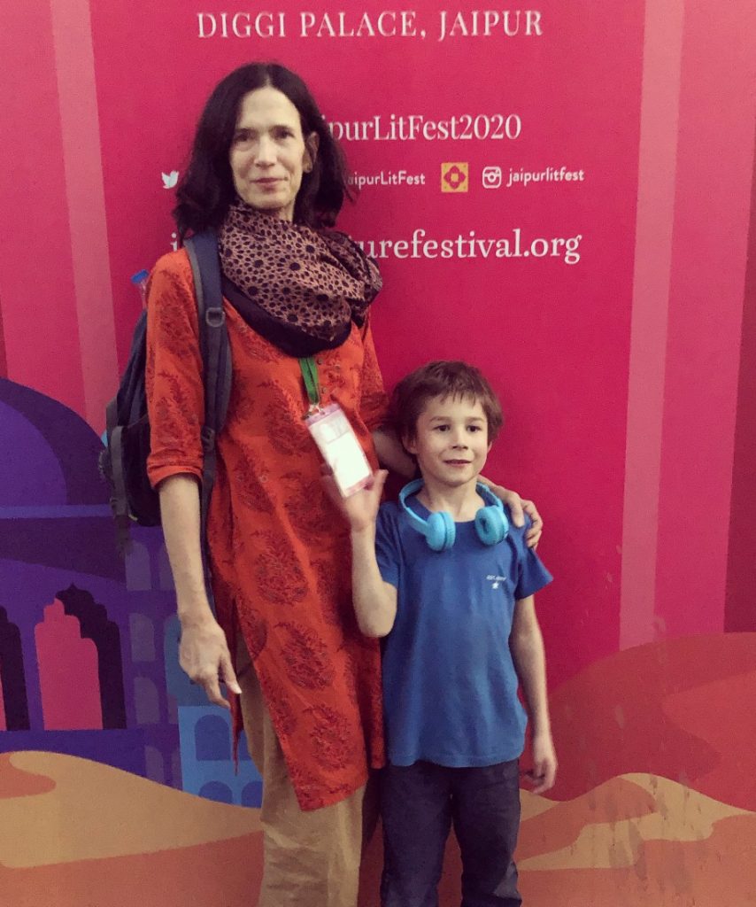 Elizabeth Kadetsky in front of a sign for the Jaipur Literature Festival, wearing her press badge and standing with her young son, Alexander.