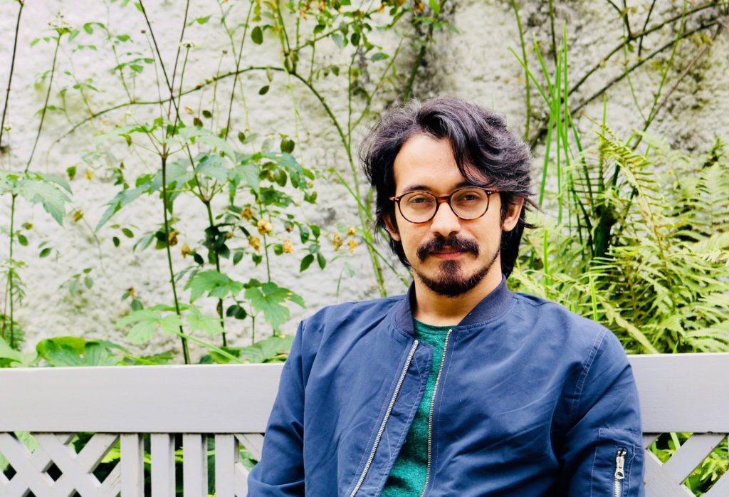 NER Poet Rohan Chhetri sits on a white bench with beard, blue jacket, and yellow flowers